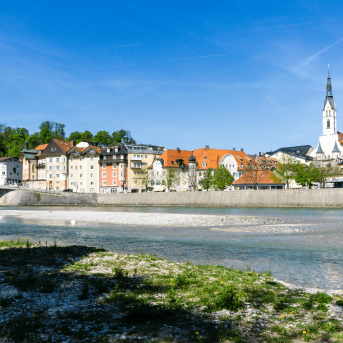 Panorama von Bad Tölz mit Blick auf die Isar, farbigen Altstadthäusern und der markanten Stadtpfarrkirche Maria Himmelfahrt an einem sonnigen Tag.