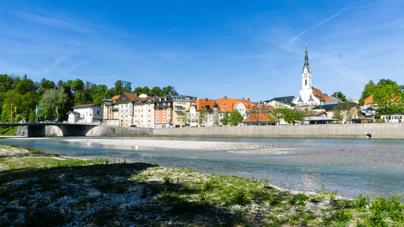 Panorama von Bad Tölz mit Blick auf die Isar, farbigen Altstadthäusern und der markanten Stadtpfarrkirche Maria Himmelfahrt an einem sonnigen Tag.